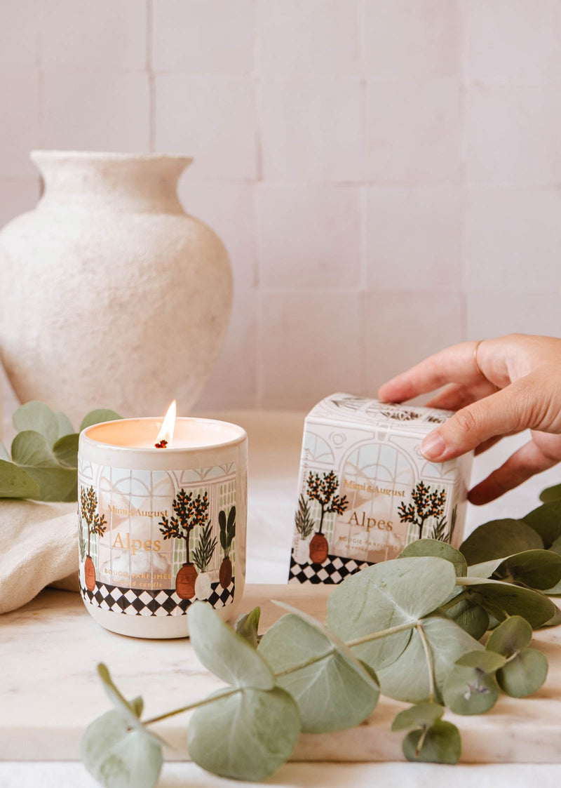 Une bougie réutilisable Alpes allumée de Mimi & August dans un récipient en céramique et sa boîte décorative sur une table ornée de feuillage vert, avec une main tenant la boîte réutilisable. Un vase blanc est placé en arrière-plan.