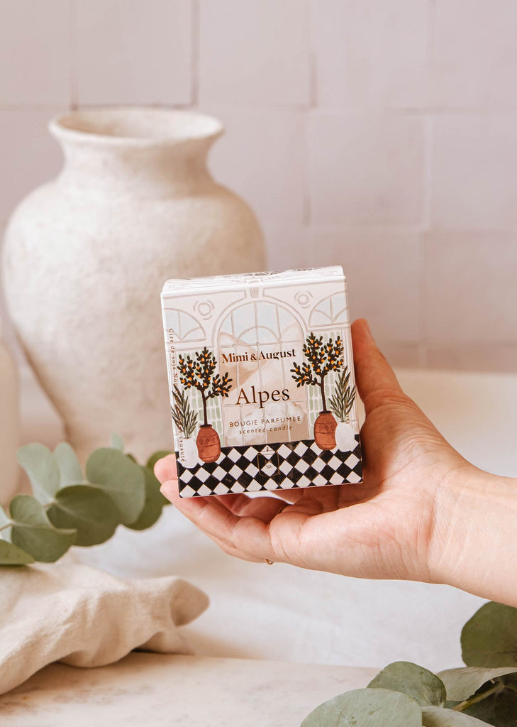 A hand holds a box of "Mimi & August Alpes - Reusable Candle" with an illustrated design. A white ceramic vase and green eucalyptus leaves are in the blurred background, hinting at the fresh scent and elegance of these reusable candles.