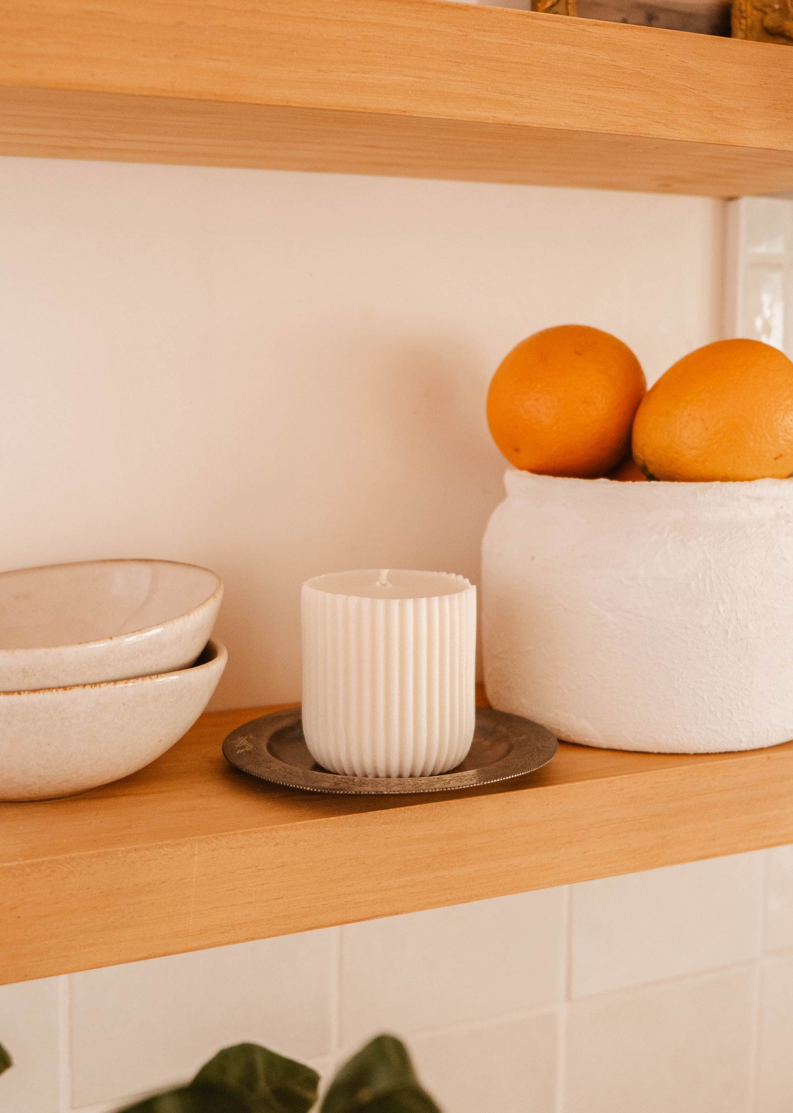 Une étagère en bois avec deux bols en céramique blanche, une bougie blanche striée - Alpes par Mimi & August sur une plaque métallique dégageant un arôme vivifiant, et trois oranges dans un récipient blanc.