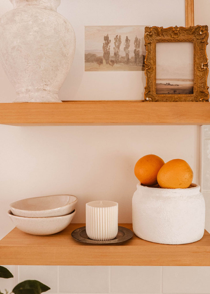 Wooden shelves holding a large vase, framed pictures, a white container with oranges, a Candle Refill - Alpes by Mimi & August on a saucer releasing an invigorating aroma of Alpes scent, and ceramic bowls.