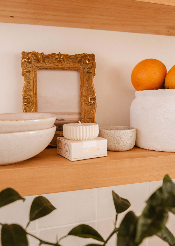 A wooden shelf with ceramic bowls, framed artwork, a white container holding oranges, and an eco-friendly Candle Refill - Alpes by Mimi & August that emits an invigorating aroma reminiscent of the Alpes. Leaves are partially visible at the bottom.