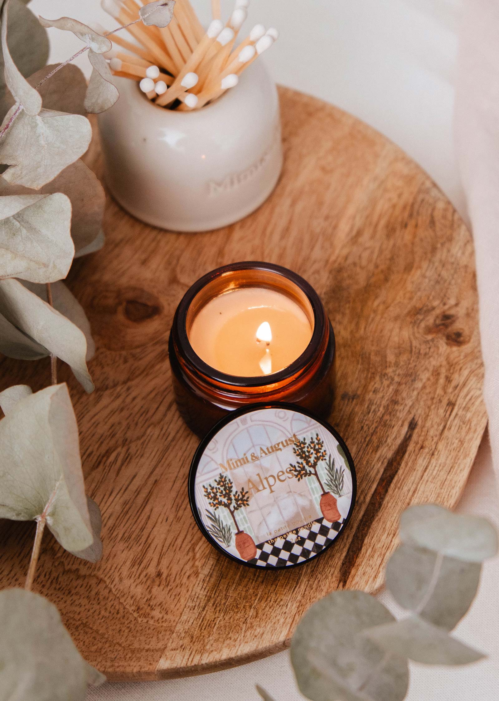 A lit Mimi & August Alpes - 2 oz reusable candle in a brown glass jar sits on a wooden tray, its soothing scent mingling with a lidded container of cotton swabs and dried eucalyptus leaves.