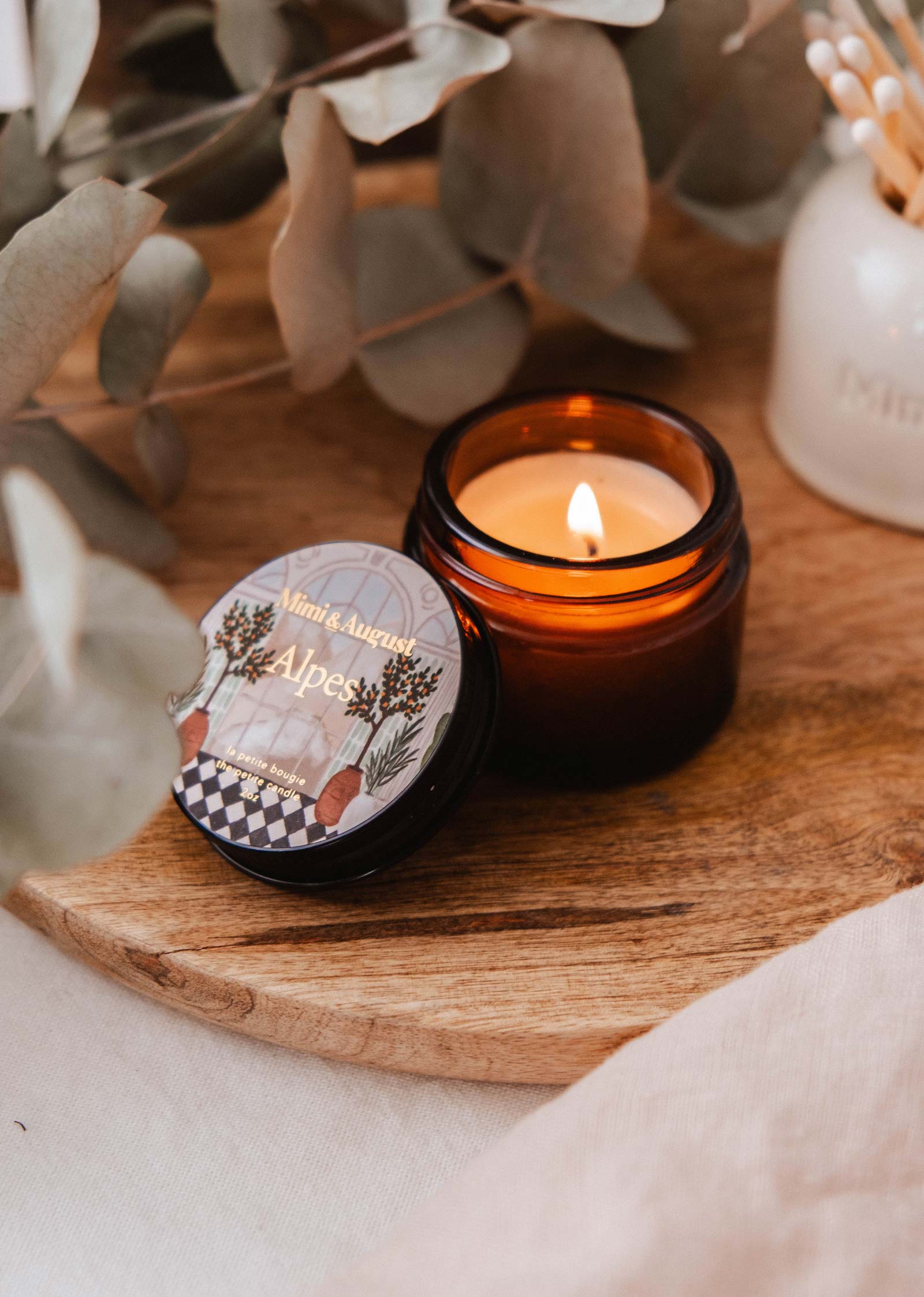 A lit brown glass candle from Mimi & August sits on a wooden tray beside a closed tin labeled "Alpes - 2 oz," surrounded by eucalyptus, evoking Alpine freshness.