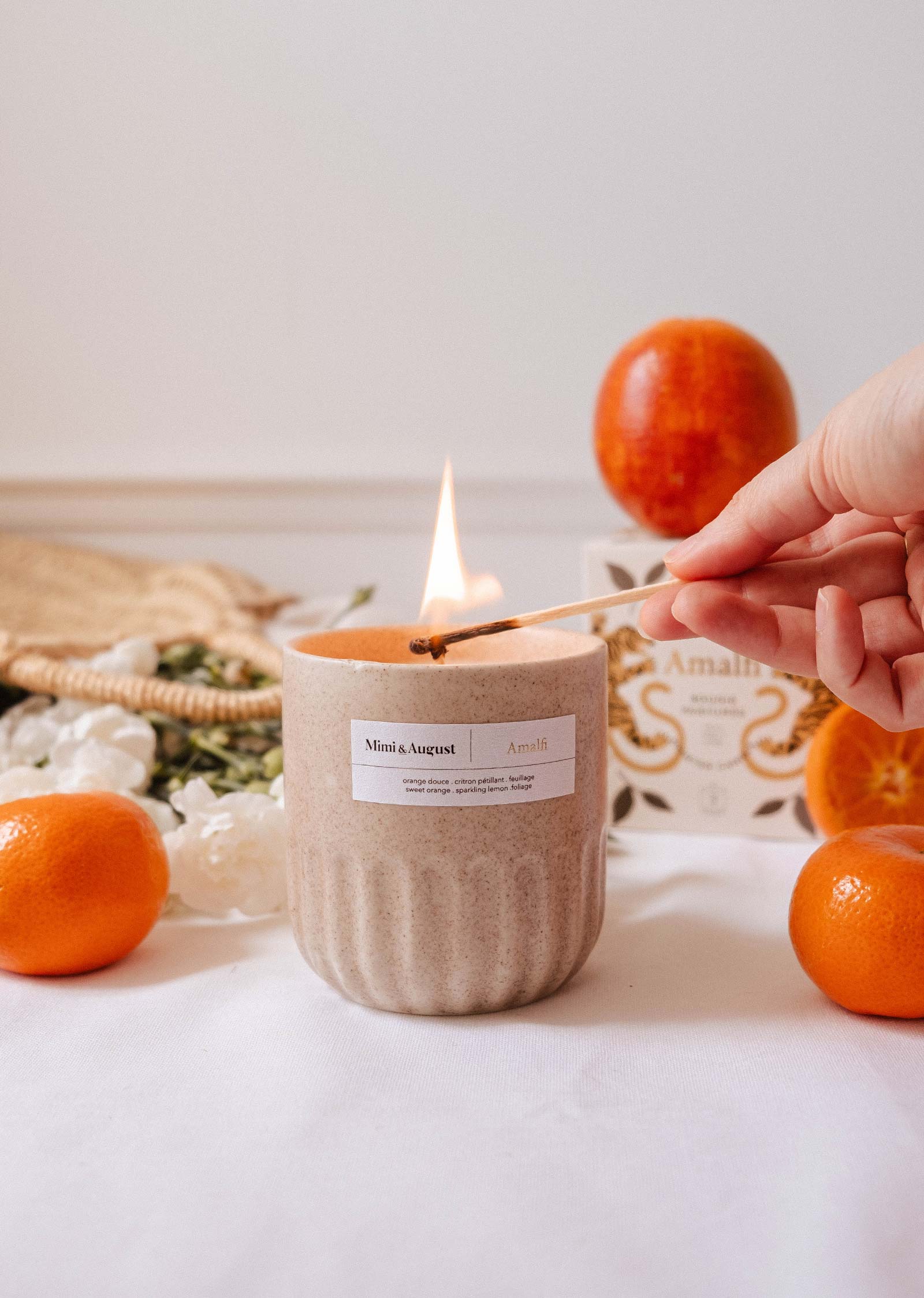 A hand lights the Amalfi - Reusable Candle by Mimi & August in a beige jar on a white surface, with oranges, a box, and tropical foliage nearby.
