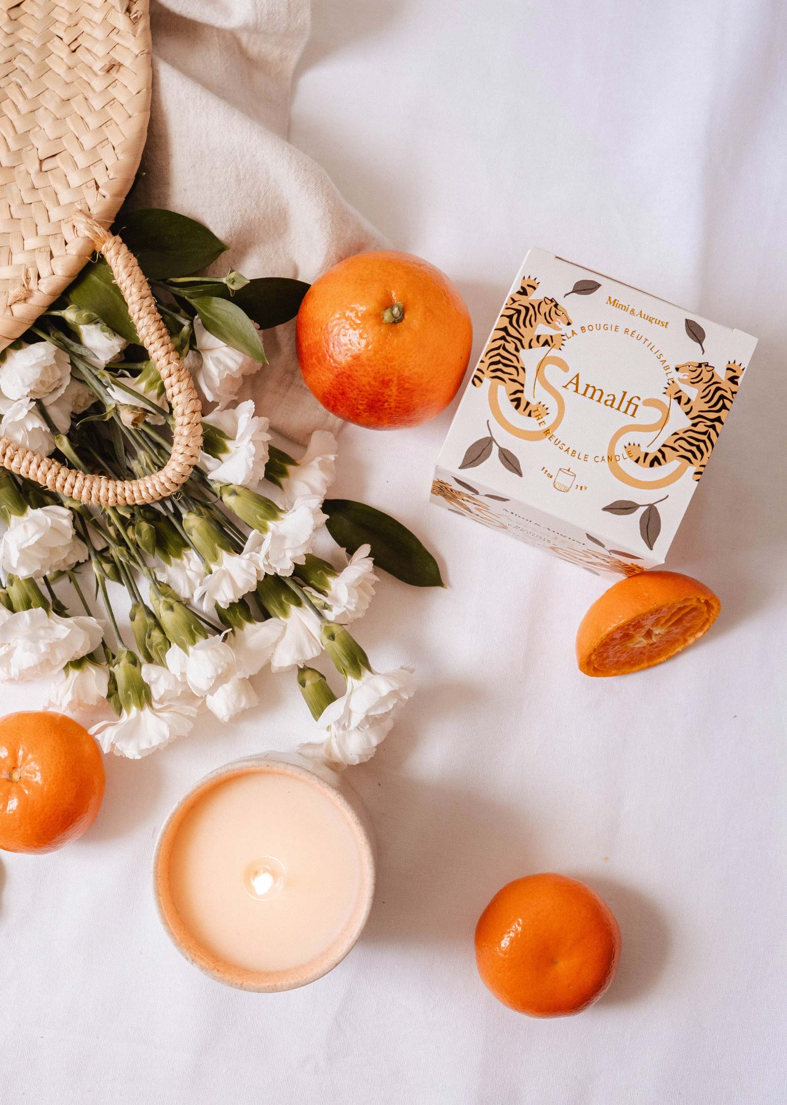 A woven bag with white flowers, oranges, and a lit Mimi & August Amalfi - Reusable Candle evokes memories of seaside holidays, all beautifully arranged on a white surface.
