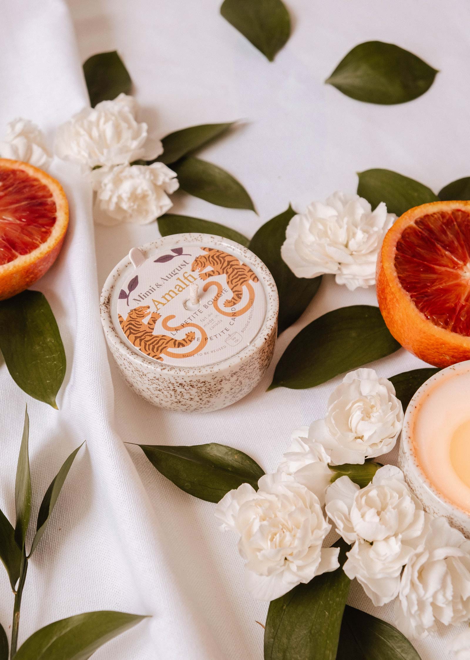 A round "Amalfi - Reusable Candle" by Mimi & August rests amid white flowers, tropical leaves, and blood orange slices on white fabric, capturing the essence of seaside holidays.