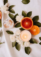 Une bougie réutilisable allumée Mimi & August Amalfi 3 oz, des tranches d'orange sanguine et d'orange, un pot rond, des fleurs blanches et un feuillage tropical luxuriant sont disposés sur une surface blanche.