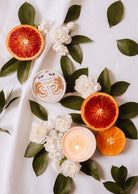 Des tranches d'oranges sanguines, des fleurs blanches, des feuilles tropicales et la bougie réutilisable Mimi & August Amalfi 3 oz sont joliment disposées sur un tissu blanc, évoquant des souvenirs de vacances au bord de la mer.