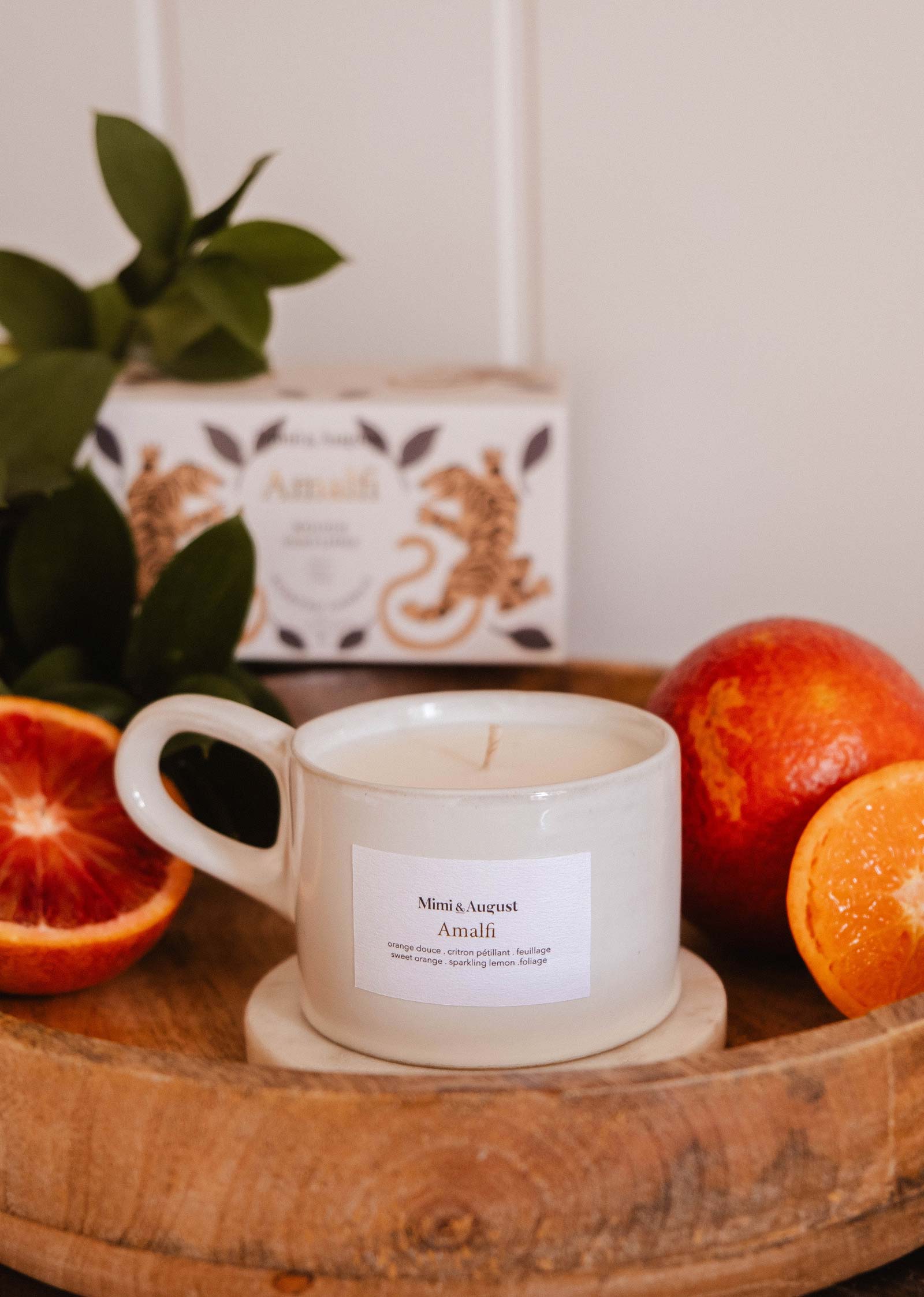 A white Mimi & August Amalfi - Reusable Candle in a cup rests on a wooden tray with blood oranges, tropical leaves, and a decorative box, capturing the seaside holiday charm.