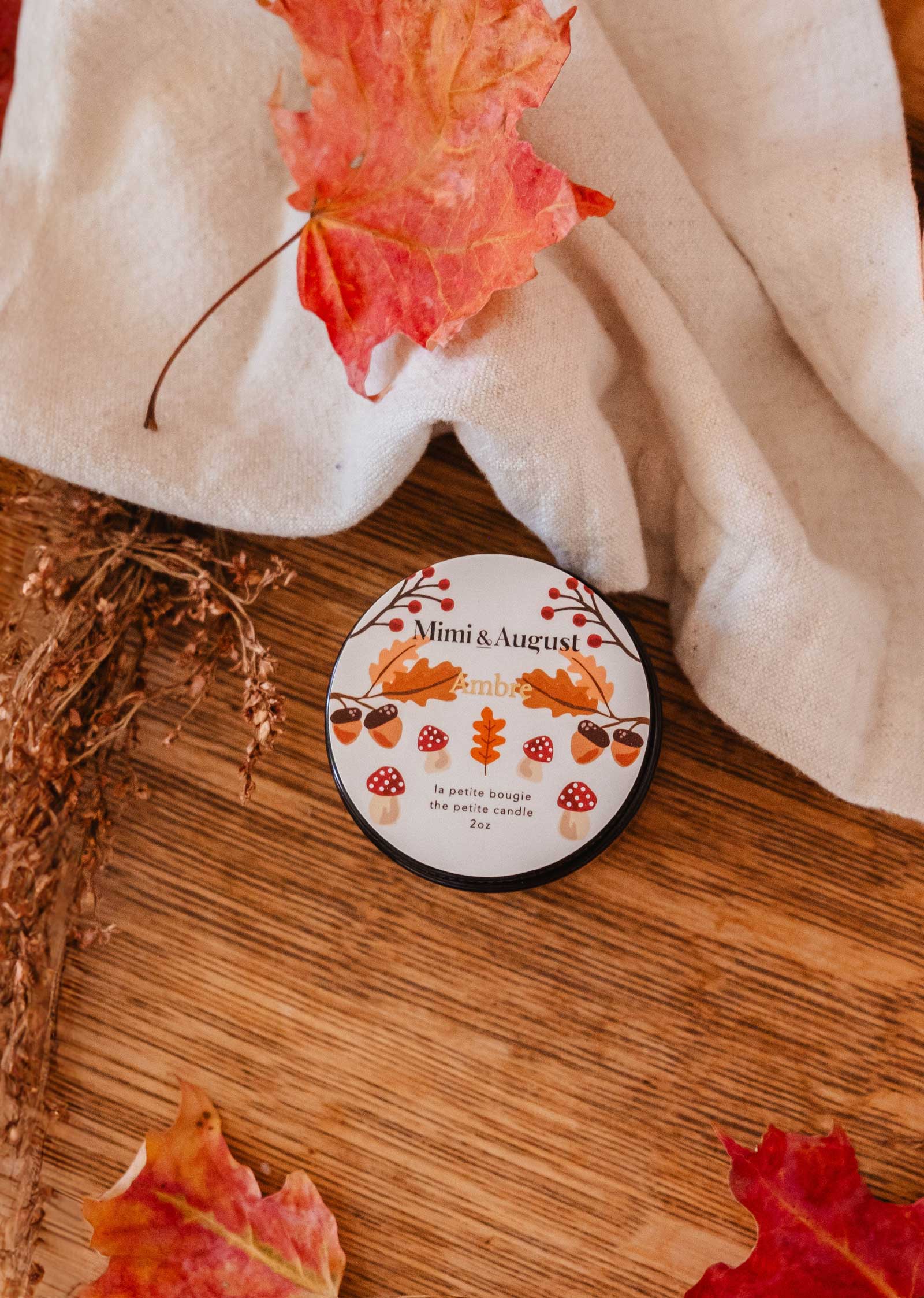 A reusable candle labeled "Ambre" by Mimi & August is placed on a wooden surface adorned with autumn leaves and a linen cloth, exuding fall coziness and a tranquil atmosphere.