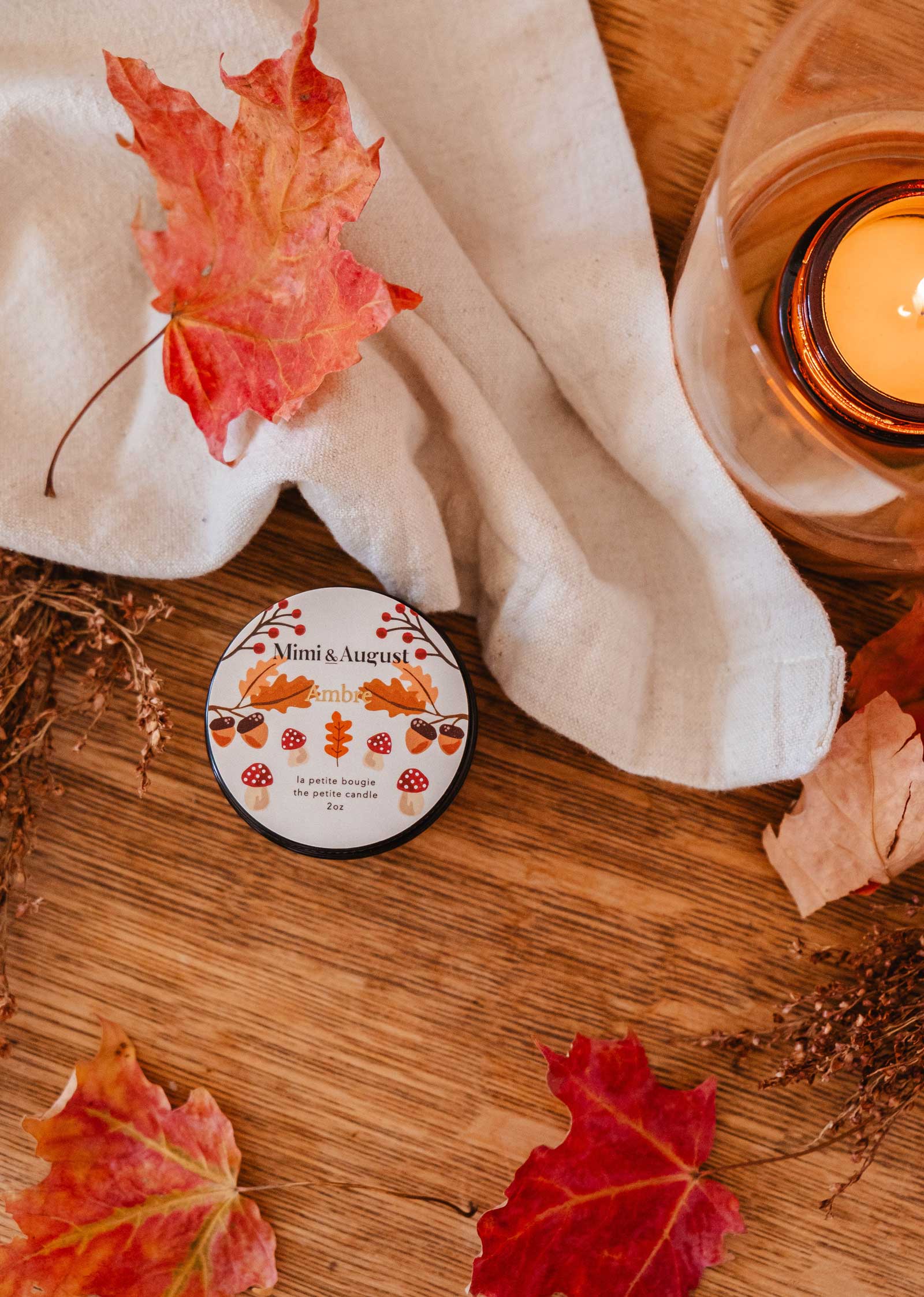 A tin labeled "Ambre - Reusable Candle" by Mimi & August rests on a wooden surface surrounded by autumn leaves, a lit candle radiating fall coziness, and a white cloth nearby, creating a tranquil atmosphere.