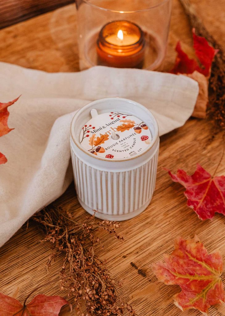 A white, ridged container labeled "Ambre" from Mimi & August sits on a wooden surface surrounded by autumn leaves and a lit orange candle in a glass holder. A beige cloth is draped nearby, enhancing the warm notes of the scene.