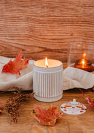 Une bougie allumée dans un support en céramique blanche nervurée repose sur une surface en bois ornée de feuilles d'automne éparses, de fleurs séchées et d'un tissu blanc plié, émettant des notes chaudes rappelant le lait de coco. À l'arrière-plan, une bougie réutilisable Ambre de Mimi & August, enfermée dans un verre, brille doucement.