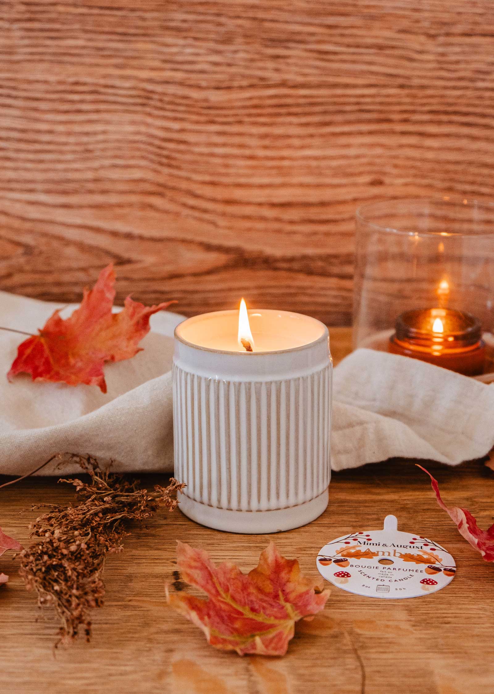 A lit candle in a white ribbed ceramic holder sits on a wooden surface adorned with scattered autumn leaves, dried flowers, and a folded white cloth, emitting warm notes reminiscent of coconut milk. In the background, an Ambre - Reusable Candle by Mimi & August encased in glass softly glows.