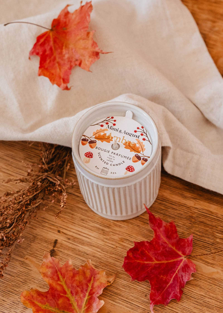 An Ambre - Reusable Candle by Mimi & August, featuring warm notes of coconut milk and a white exterior with an autumn-themed lid, sits on a wooden surface surrounded by red and orange maple leaves and a beige cloth. This candle adds a cozy touch to any space.