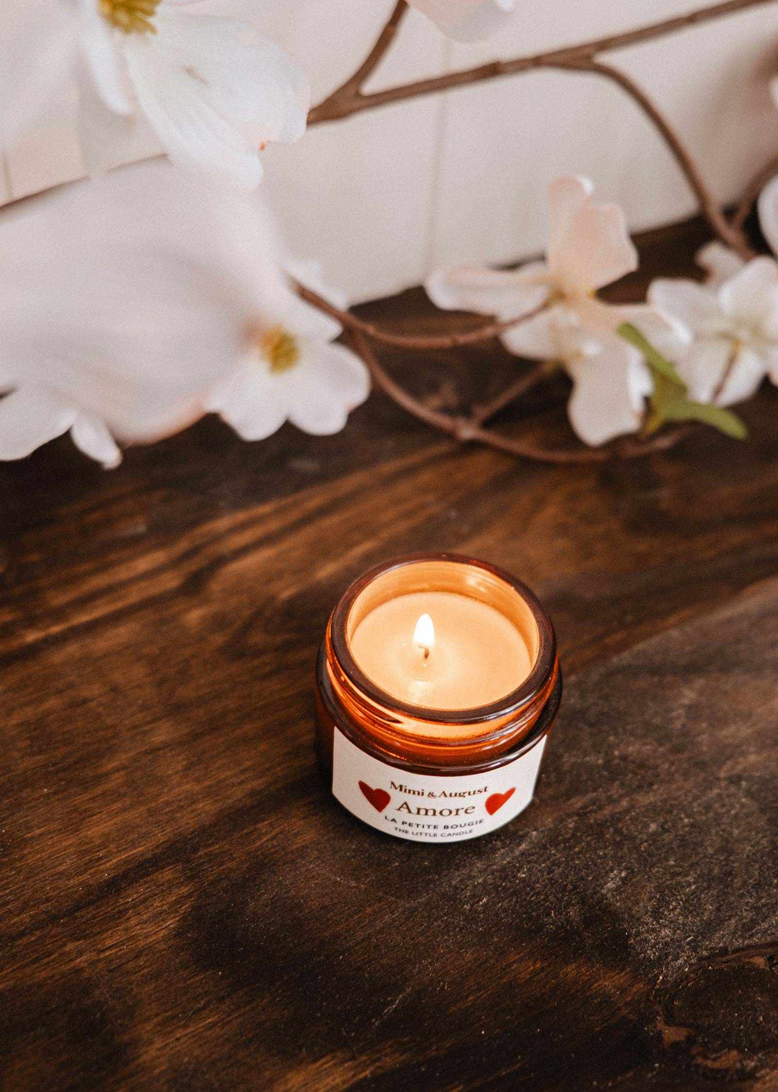 Une bougie de soya Amore 2 oz de Mimi & August dans un pot en verre brun repose sur une surface en bois, avec des fleurs blanches en arrière-plan.