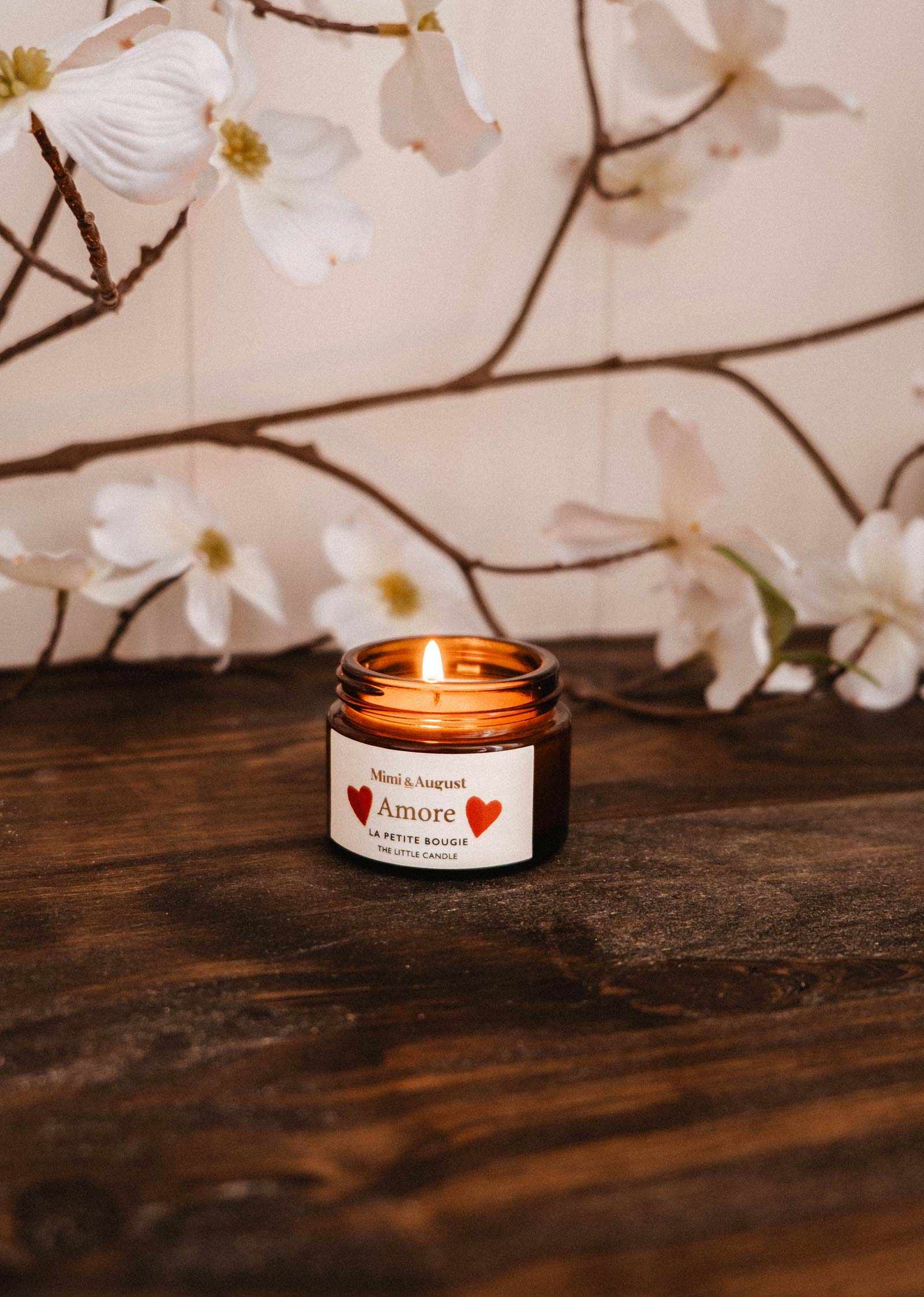 La bougie de soya Amore 2 oz de Mimi & August, dans un petit contenant en verre ambré, brûle sur du bois avec des fleurs blanches et des branches en arrière-plan — une bougie de soya romantique dans un pot en verre réutilisable.