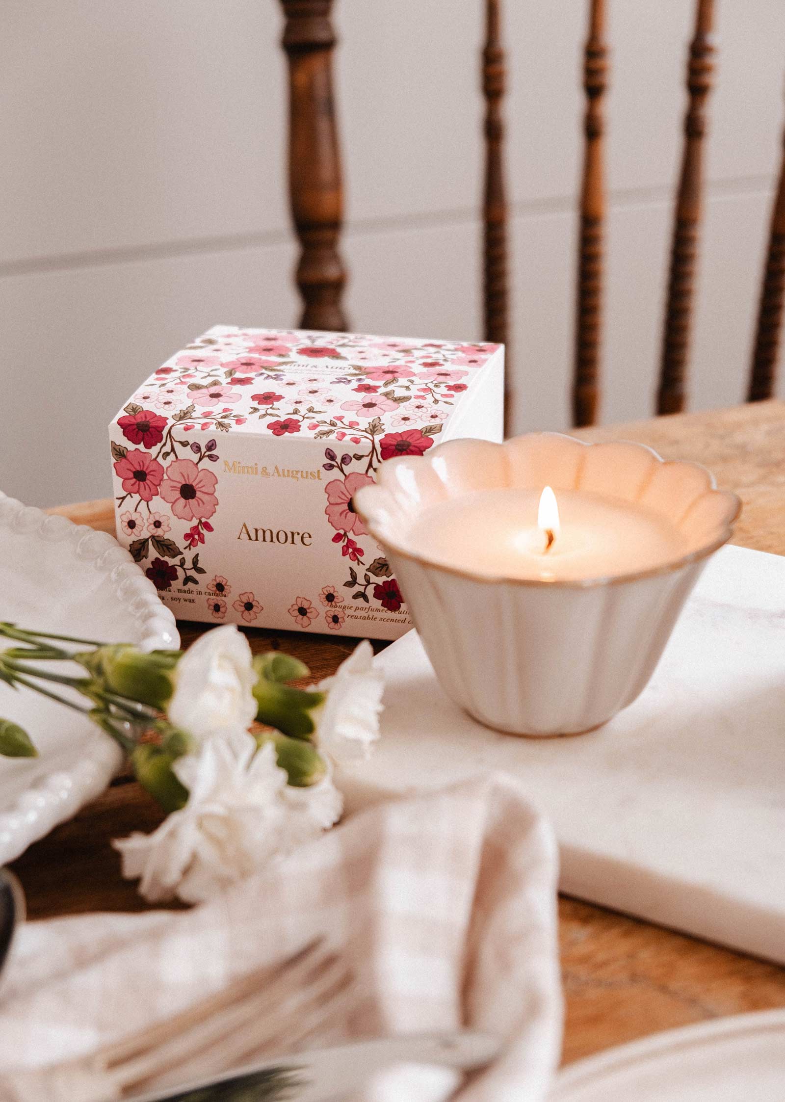 A Mimi & August Amore Reusable Candle in a scalloped white holder rests beside a floral "Amore" box and a small bouquet of white flowers.