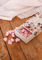 An open floral box on wood spills pink heart-shaped Amore Scented Wax Melts by Mimi & August, with white fabric in the background, evoking a romantic fragrance.