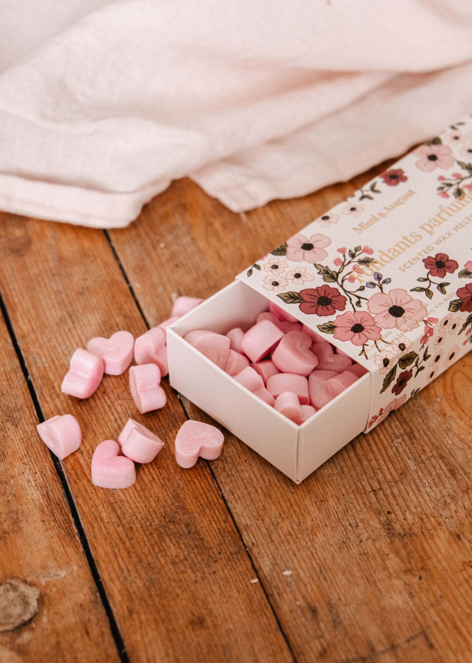 An open floral box on wood spills pink heart-shaped Amore Scented Wax Melts by Mimi & August, with white fabric in the background, evoking a romantic fragrance.