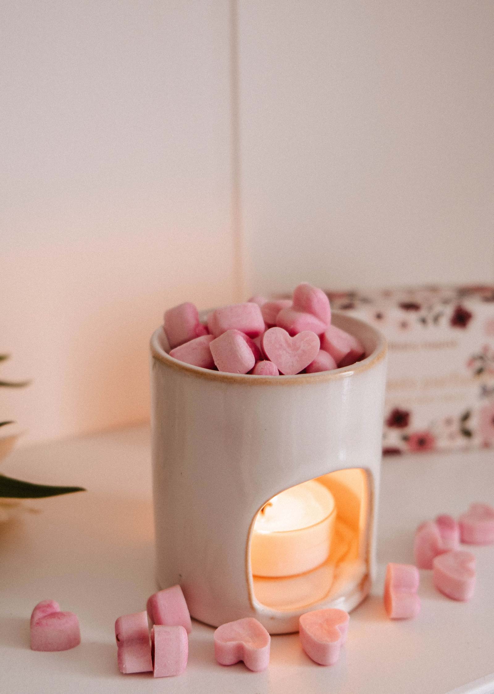 A white ceramic wax warmer holds pink, heart-shaped Amore Scented Wax Melts by Mimi & August, radiating a romantic aroma with a lit tealight candle inside and extra heart-shaped melts scattered nearby.
