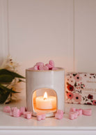 A white ceramic candle holder with a lit tealight, filled with Mimi & August’s Amore Scented Wax Melts in pink heart shapes, sits on a white surface with scattered melts and a floral box in the background, creating a romantic aroma.