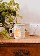 On a wooden table, a small lit candle rests in a speckled holder, accompanied by a green plant and an essential oil bottle nearby. In the background, there is an Aromatic Ceramic Diffuser from Mimi & August.