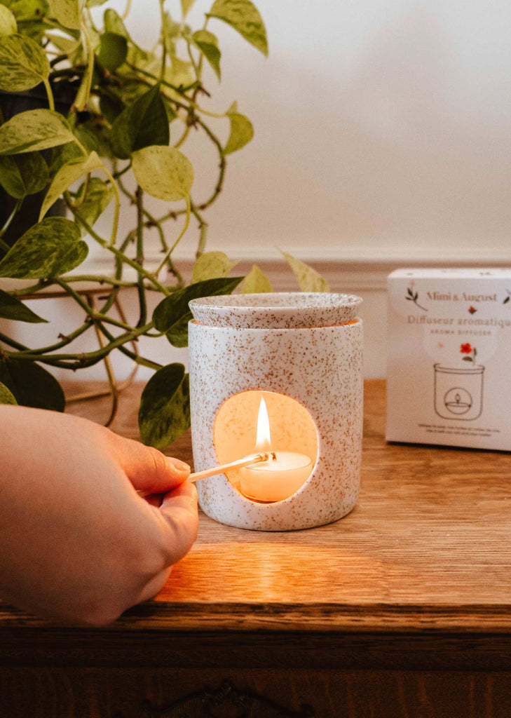 Une main allume une bougie à l'intérieur du diffuseur aromatique en céramique de Mimi & August sur une surface en bois, avec une plante feuillue et une boîte en arrière-plan.