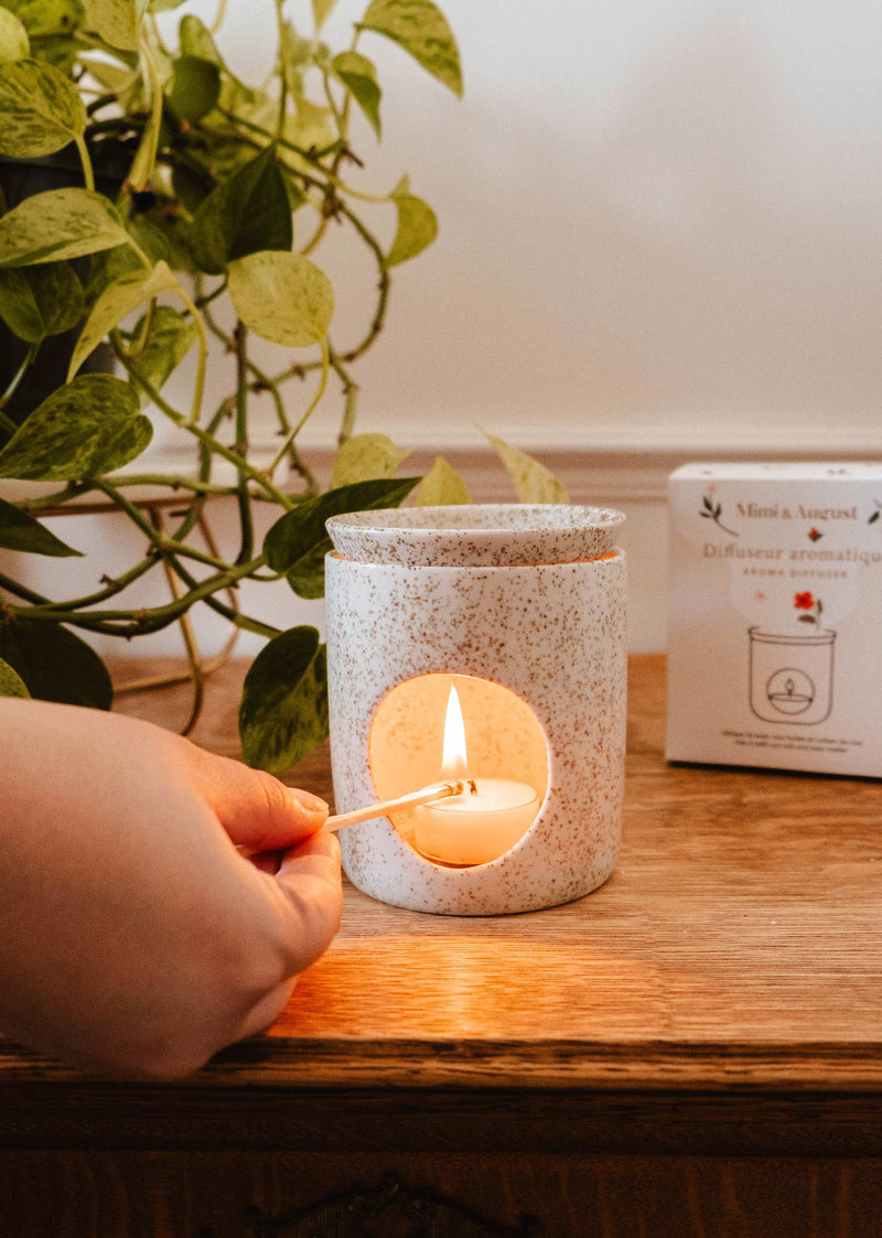 Une main allume une bougie à l'intérieur du diffuseur aromatique en céramique de Mimi & August sur une surface en bois, avec une plante feuillue et une boîte en arrière-plan.