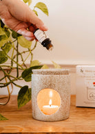 A hand pours essential oil into the Aromatic Ceramic Diffuser by Mimi & August, which has a lit tealight candle inside and is placed on a wooden surface, with a green potted plant in the background.