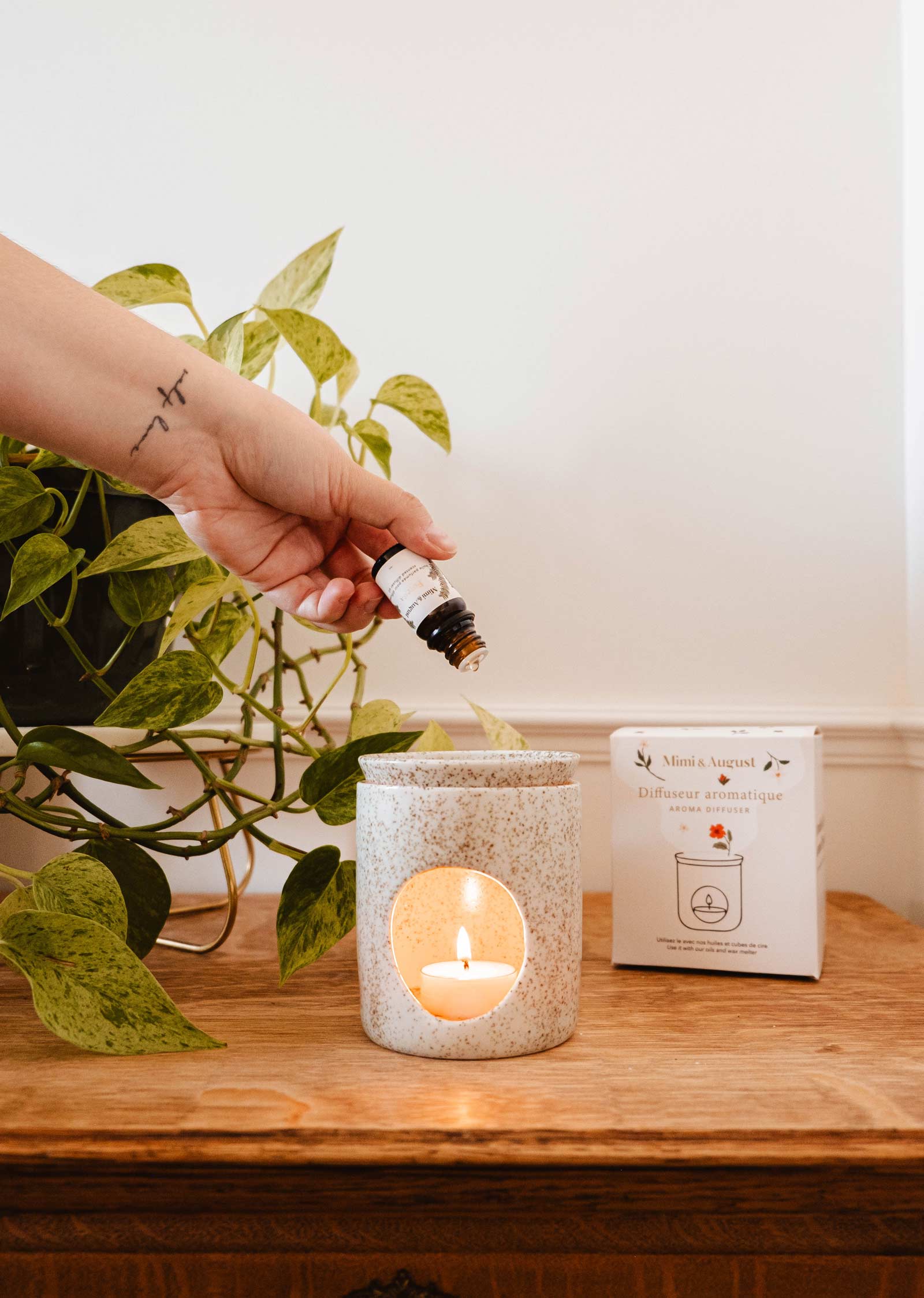 A hand pours essential oil into the Aromatic Ceramic Diffuser by Mimi & August, which has a candle lit inside. In the background, there's a green plant and a product box.