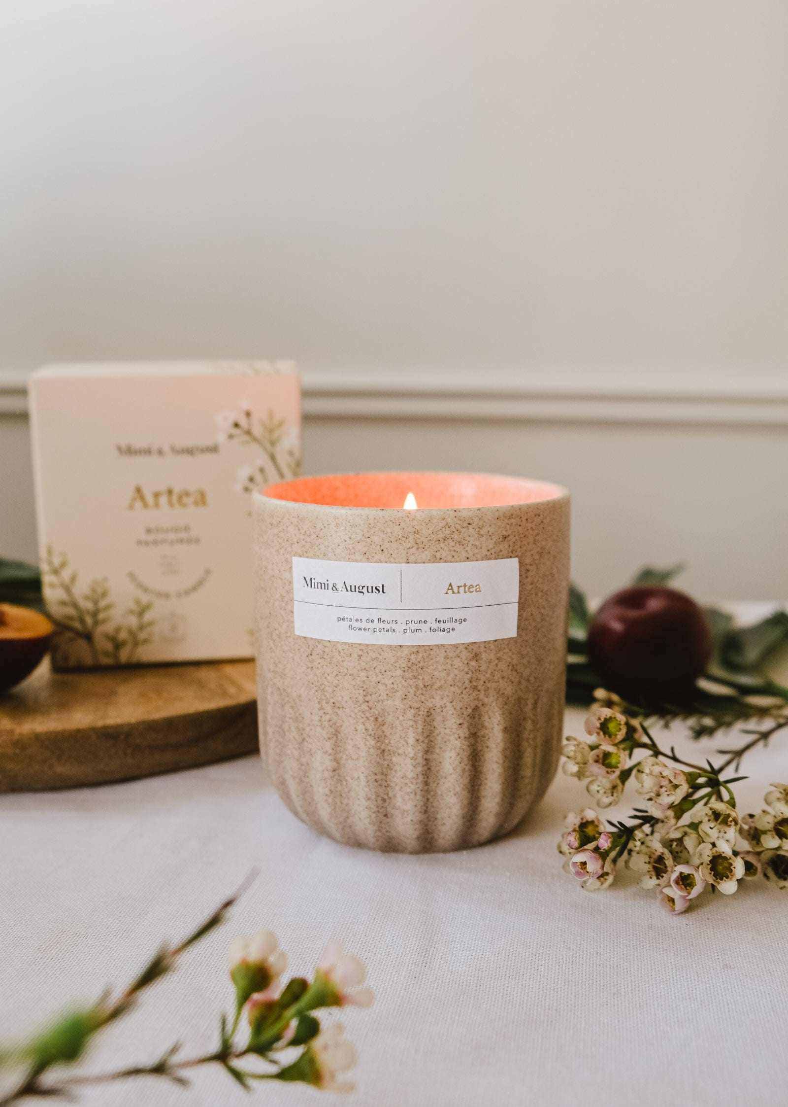 A beige floral Mimi & August Artea – Reusable Soy Candle is lit on a table with small flowers, greenery, and its box in the background, adding charm and sustainability to any space.