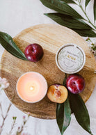 A lit Mimi & August Artea – Reusable Soy Candle in a ceramic container, plums, a halved plum, and the candle’s tin are arranged on a round wooden board with green leaves.
