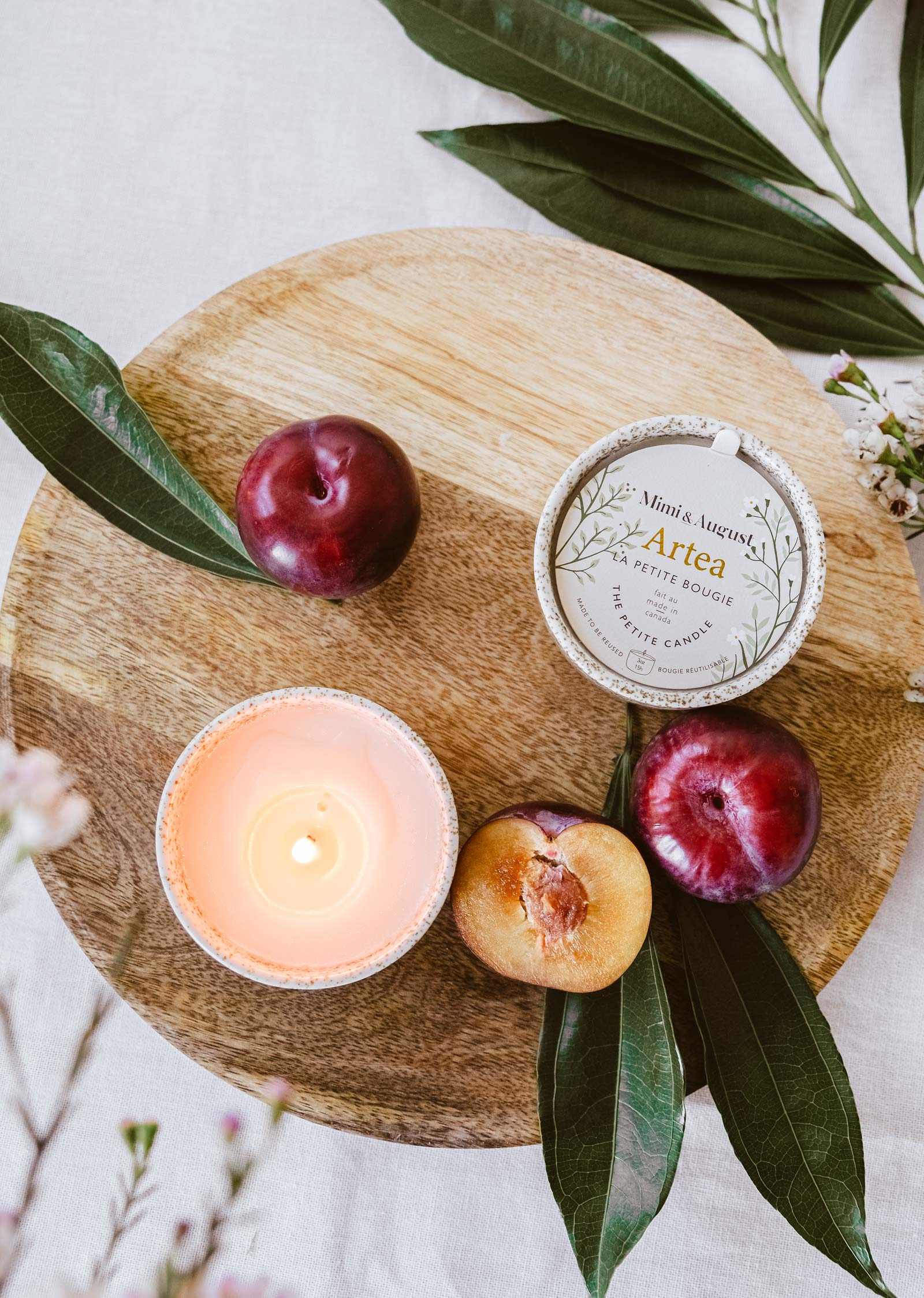 A lit Mimi & August Artea – Reusable Soy Candle in a ceramic container, plums, a halved plum, and the candle’s tin are arranged on a round wooden board with green leaves.