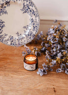 La bougie de soya Azalée 2 oz de Mimi & August, coulée à la main à Montréal et présentée dans un pot en verre, repose sur une surface en bois à côté de fleurs violettes et d'une assiette blanche aux motifs floraux bleus.