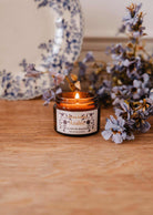 La bougie de soya Azalée 2 oz de Mimi & August, coulée à la main à Montréal, repose dans un pot en verre brun sur du bois à côté de fleurs violettes et d'une assiette décorative, apportant chaleur et élégance à tout espace.