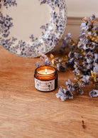 La bougie de soya Azalée 2 oz de Mimi & August, coulée à la main à Montréal, repose dans un pot en verre brun sur du bois à côté de fleurs violet pâle et d'une assiette décorative bleue et blanche.