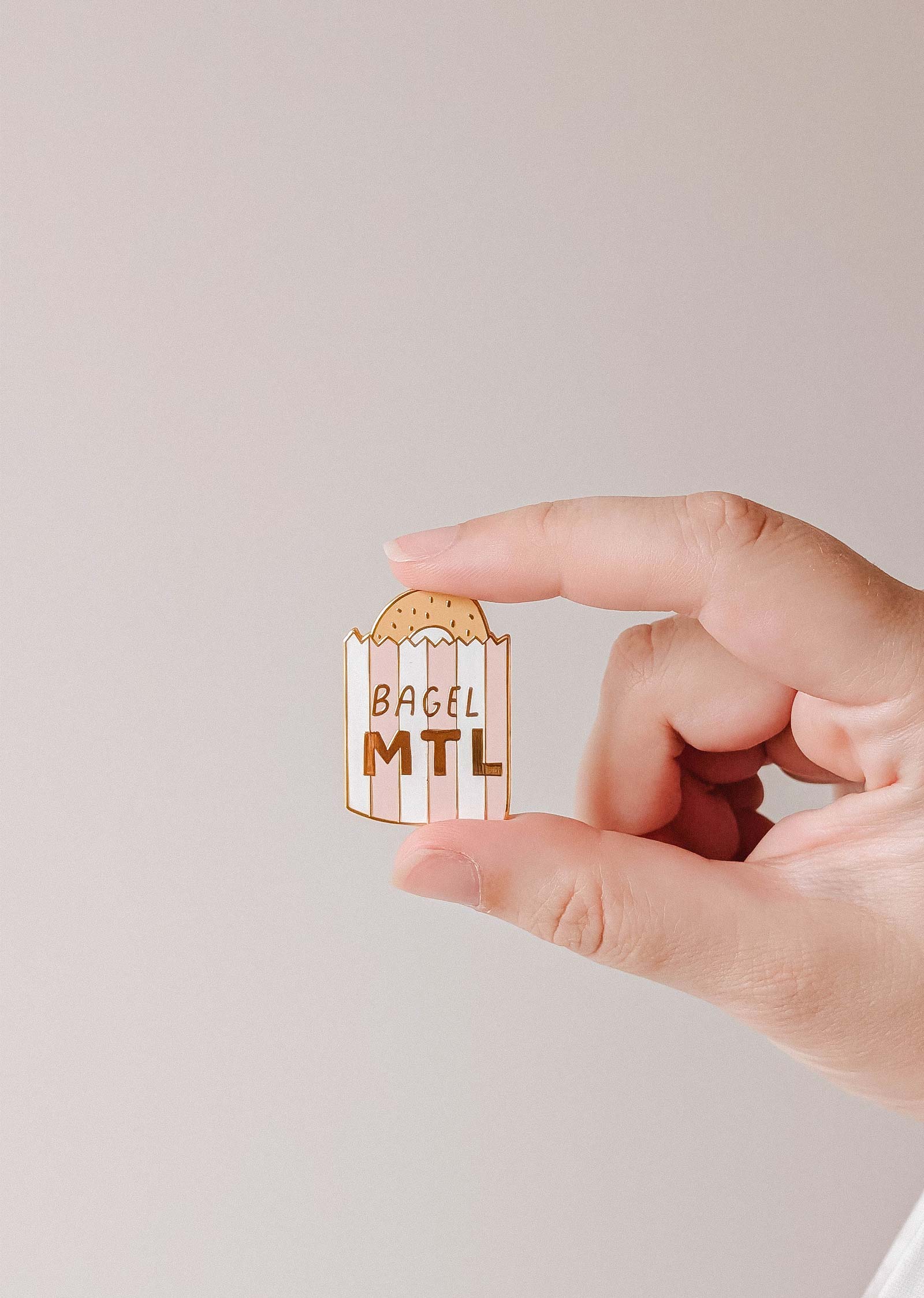 A hand holds a Bagel MTL Enamel Pin by Mimi & August, shaped like a Montréal bagel bag with "BAGEL MTL" on it—an ideal accessory for foodies and bagel lovers.