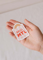 A hand holds the Bagel MTL - Vinyl Sticker by Mimi & August, showcasing a Montréal icon—a bagel nestled in a paper bag—on durable vinyl.