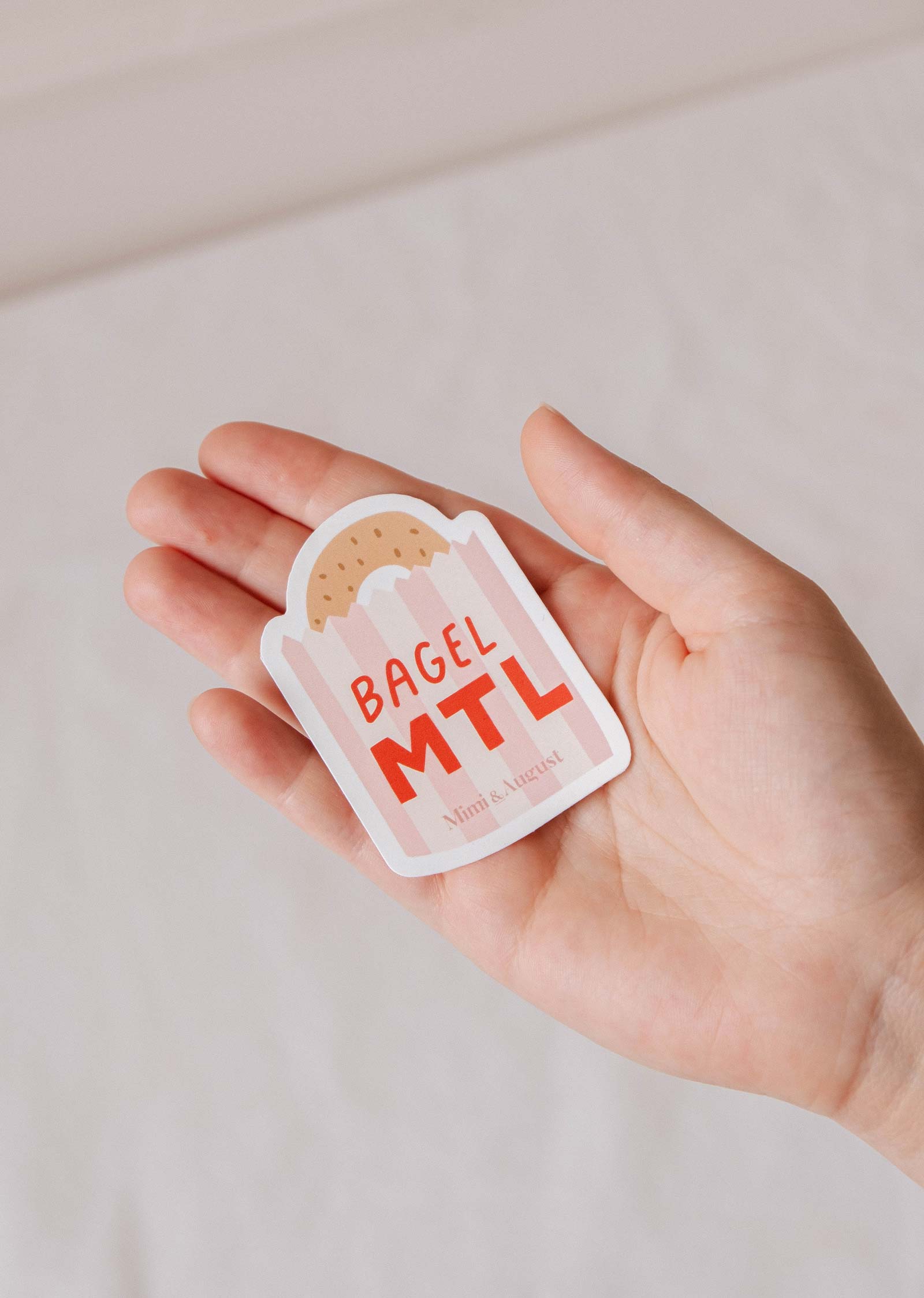 A hand holds the Bagel MTL - Vinyl Sticker by Mimi & August, showcasing a Montréal icon—a bagel nestled in a paper bag—on durable vinyl.