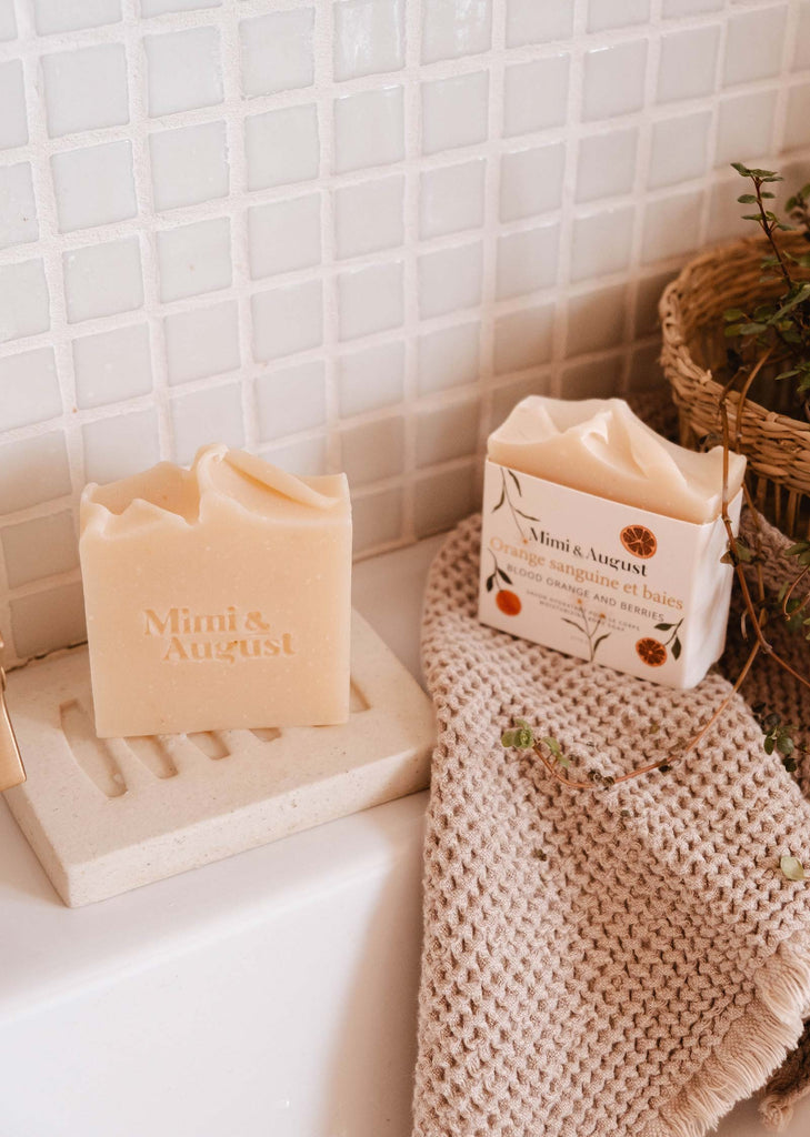 A bar of hydrating Mimi & August Bar Soap Blood Orange & Berries and its packaging on a bathroom counter with a waffle-weave towel and a basket in the background.