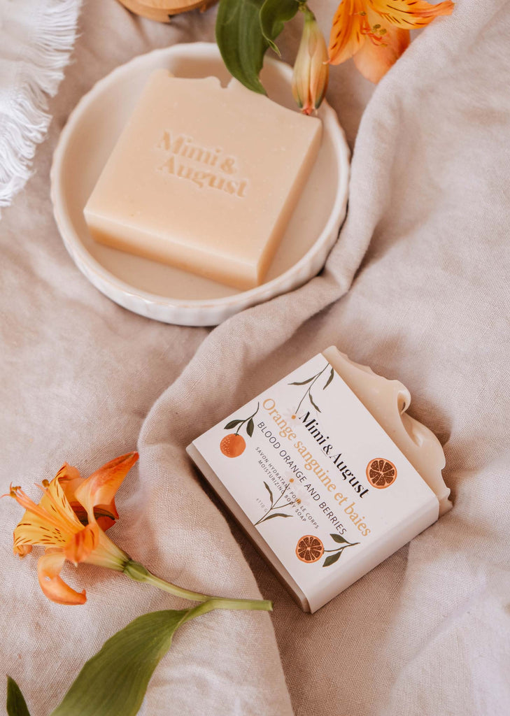 Arrangement esthétique du savon en barre hydratant orange sanguine et baies de Mimi & August avec des décorations florales sur une surface en tissu doux.