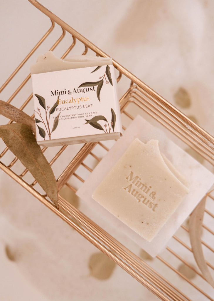 Mimi & August Eucalyptus Bar Soap alongside its packaging on a wire rack with a eucalyptus leaf, known for its natural stress reliever qualities.