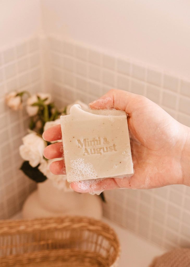A person holds a bar of Mimi & August Eucalyptus Bar Soap over a bathtub with a floral arrangement and a woven basket in the background.