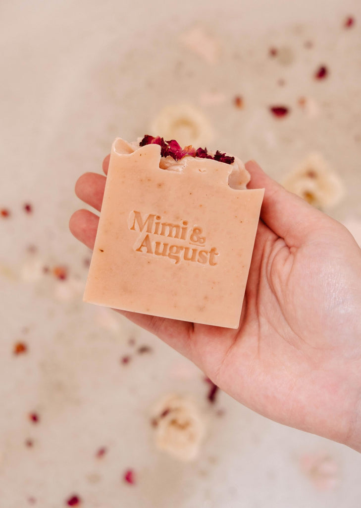 Hand holding a Mimi & August Sweet Rose Bar Soap with "mimi & august" embossed on it, surrounded by scattered petals.