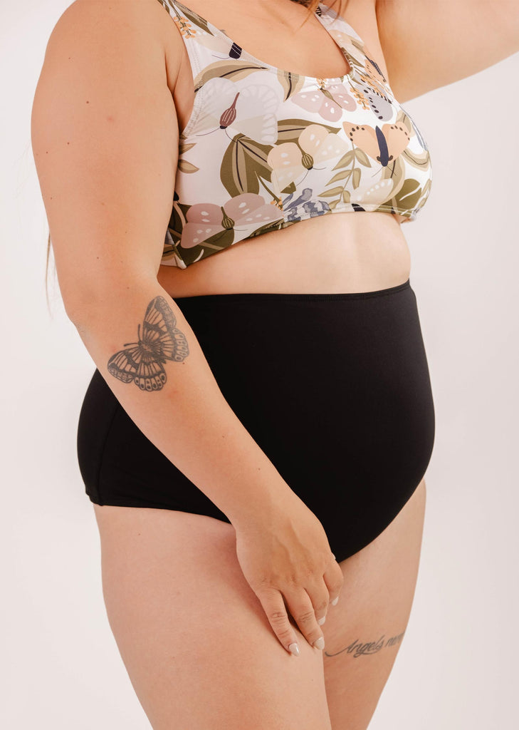 A person wearing a floral bikini top and Bermudes Black extra high waisted bikini bottom by Mimi & August shows off a tattoo of a butterfly on their upper arm, showcasing a flattering silhouette.