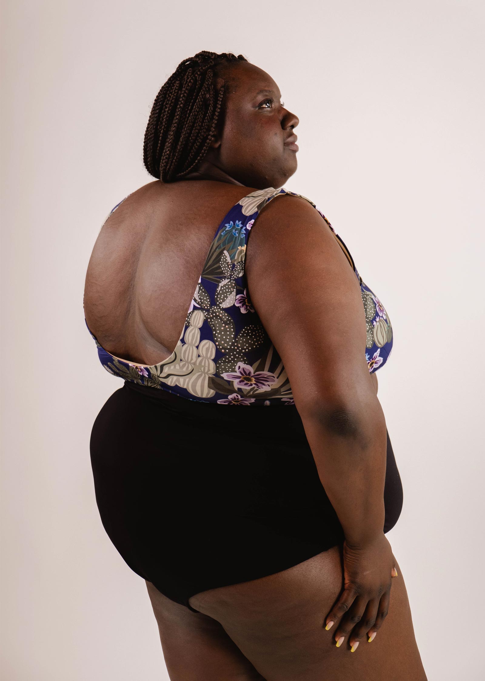 A person with braided hair wearing a floral-patterned swimsuit, featuring a Bermudes Black extra high waisted bikini bottom by Mimi & August, poses against a plain background.