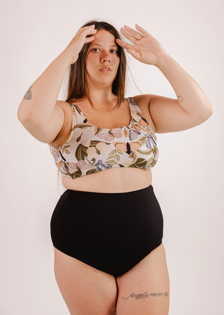 A woman with long hair wearing a floral bikini top and flattering, Mimi & August Bermudes Black extra high waisted bikini bottom stands against a neutral background, her hands raised near her head.