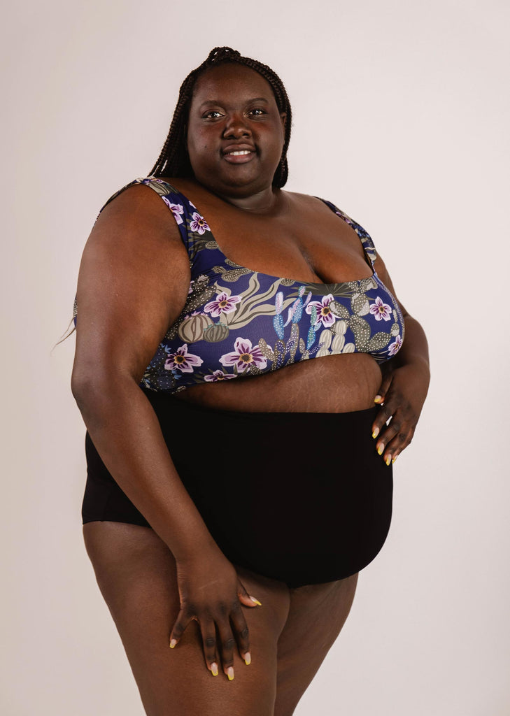 Une personne portant un haut de bain à motifs floraux et un bas de bikini taille haute extra Bermudes Noir de Mimi & August, debout sur un fond uni, créant une silhouette flatteuse.