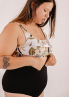 A woman with long brown hair adjusts her floral print top. She is wearing a flattering silhouette with Mimi & August Bermudes Black extra high-waisted bikini bottoms and has a tattoo of flowers on her forearm.